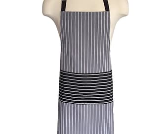 May include: A gray and white striped apron with a black waistband and neck strap. The apron is displayed on a mannequin. The apron is designed for kitchen or craft use.