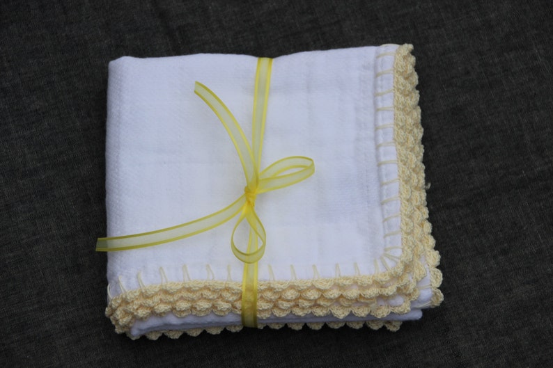 May include: A stack of white, square cloths with a crocheted cream border. The cloths are tied with a yellow ribbon. The background is a dark, plain colour.