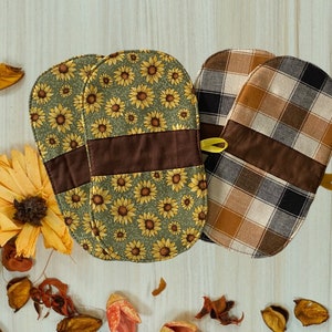 May include: Two oven mitts with a sunflower pattern and a brown band, and two oven mitts with a plaid pattern and a brown band. The mitts have yellow loops for hanging. The background includes dried flowers and leaves.