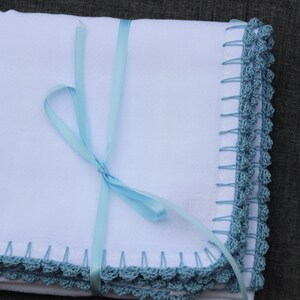 May include: A white blanket with a blue crocheted edge and a light blue ribbon tied around it. The blanket has a handmade look, possibly a baby blanket or similar item. The crocheted border is in a blue-green hue.