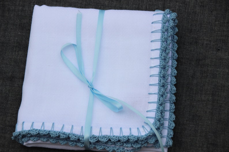May include: A white blanket with a blue crocheted edge and a light blue ribbon tied around it. The blanket has a handmade look, possibly a baby blanket or similar item. The crocheted border is in a blue-green hue.