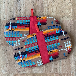 May include: Two oval pot holders with a Southwestern pattern in brown, blue, red, and orange. Each has a solid red centre panel and a small red loop for hanging. The pot holders are designed for kitchen use.
