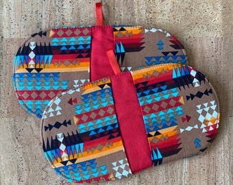 May include: Two oval-shaped pot holders with a Southwestern-inspired pattern in shades of brown, blue, red, and orange. Each pot holder has a solid red center panel and a small red loop for hanging.