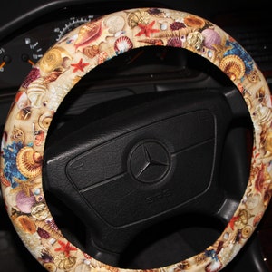 May include: A beige steering wheel cover adorned with a seashell pattern, including starfish and various shells in shades of tan, blue, and red. The cover is fitted on a black steering wheel, enhancing the interior of a vehicle.