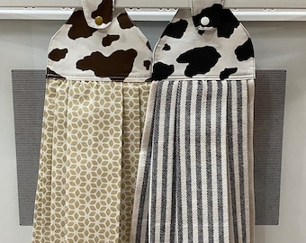 May include: Two hanging kitchen towels. One features a cow print in brown and white, the other has black and white stripes. The tops have a button closure. The towel on the left has a tan floral pattern.