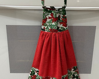 May include: A red kitchen towel with a Christmas-themed pattern of bows and holly, hanging on a white oven. The top of the towel and the bottom border feature the festive design. A sign above the oven reads "KITCHEN CLOSED."