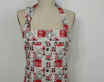 Puede incluir: Un delantal blanco con un estampado rojo y negro con temática de cocina. El delantal se exhibe en un maniquí y presenta un diseño repetido de utensilios de cocina, alimentos y electrodomésticos. El delantal tiene cuello halter y un babero cuadrado.
