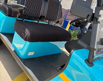 May include: A bright turquoise golf cart featuring black seats with red diamond-patterned stitching. The vehicle includes a black textured floor mat, a black roof, side mirrors, and a black fabric seat cover on the front bench seat.