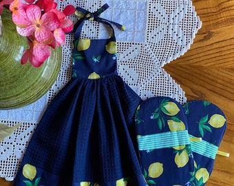 May include: A navy blue kitchen towel and matching oven mitts set. The towel has a lemon print border and a waffle weave body. The oven mitts have a lemon print and a light green stripe. The set is displayed on a white lace doily.