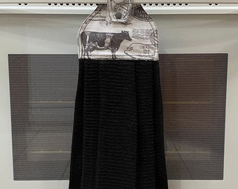 May include: A black kitchen towel with a textured pattern hangs from an oven handle. The top portion features a cream-colored fabric with a cow illustration and text. A sign that says "Kitchen Closed" is visible in the background.