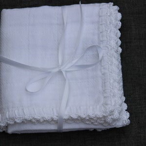 May include: A folded white blanket with a textured surface and a delicate, crocheted lace trim. The blanket is tied with a white ribbon, forming a bow. The item is presented on a dark background.