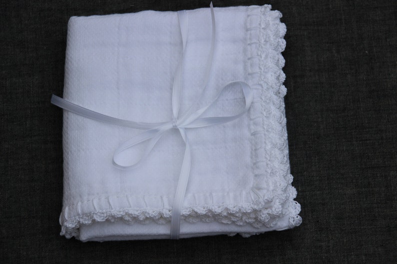 May include: A folded white blanket with a textured surface and a delicate, crocheted lace trim. The blanket is tied with a white ribbon, forming a bow. The item is presented on a dark background.