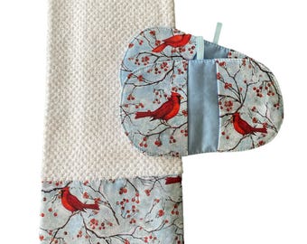 May include: A white kitchen towel and matching pot holders. The towel has a textured weave with a patterned border featuring red cardinals perched on branches with red berries against a light blue background. The pot holders have the same pattern.