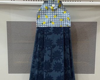 May include: A blue and white kitchen towel with a lemon pattern and a floral design. The top portion features a blue and white gingham check with yellow lemons. The bottom portion is a darker blue with a floral pattern.