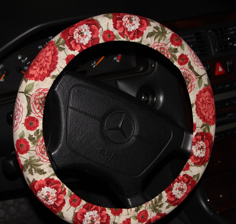 May include: A floral steering wheel cover with a pattern of red and white peony flowers and green leaves on a beige fabric. The cover is fitted on a black steering wheel with the Mercedes-Benz emblem.