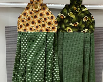 May include: Two hanging kitchen towels. One features a sunflower pattern on a tan background, the other has an avocado pattern. Both have a green towel base. Each towel has a snap closure for hanging.