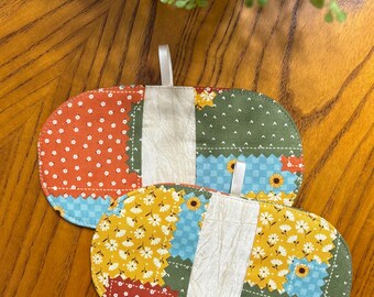 May include: Two oval-shaped, quilted pot holders with a patchwork design. One features orange polka dots, while the other has yellow daisy patterns. Both have a white ribbon loop for hanging.