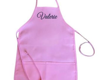 May include: A light pink apron with the name "Valerie" in cursive script. The apron has two front pockets and a neck loop. The waist ties are visible on the sides. The apron is made of a smooth, solid-colored fabric.