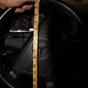 May include: Black leather steering wheel with a Mercedes-Benz logo. A wooden ruler measures the steering wheel's diameter, showing approximately 40 cm. The car's dashboard and other controls are visible in the background.
