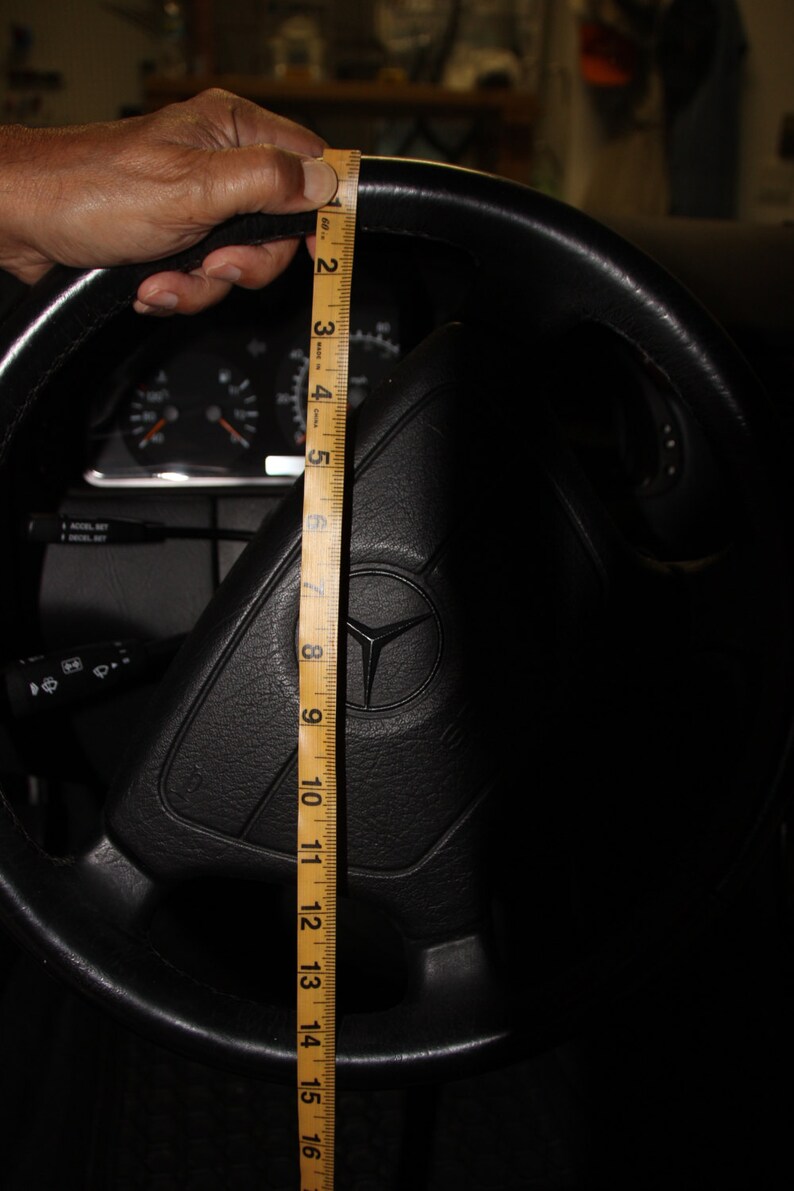 May include: Black leather steering wheel with a Mercedes-Benz logo. A wooden ruler measures the steering wheel's diameter, showing approximately 40 cm. The car's dashboard and other controls are visible in the background.