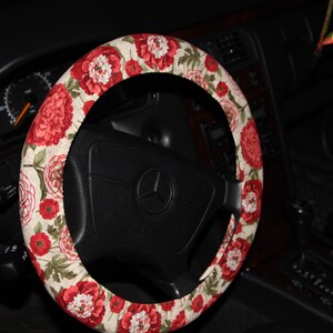 May include: A steering wheel cover with a floral design of red peonies on a cream fabric. The cover is fitted to a black steering wheel with a silver emblem. The car's interior, including the dashboard, is visible.
