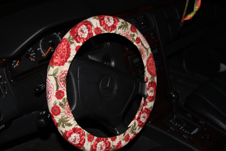 May include: A steering wheel cover with a floral design of red peonies on a cream fabric. The cover is fitted to a black steering wheel with a silver emblem. The car's interior, including the dashboard, is visible.