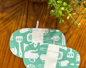 May include: Two oval-shaped, teal pot holders with white kitchen utensil patterns. Each pot holder has a white fabric band and a small white loop for hanging. The pot holders are on a wooden surface.
