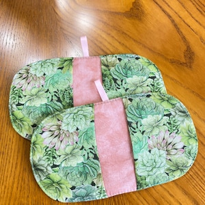 May include: Two oval-shaped pot holders with a succulent plant pattern in shades of green and pink. Each pot holder has a central pink panel and a small pink loop for hanging. The pot holders are on a wooden surface.