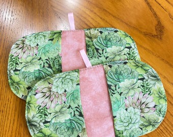 May include: Two oval-shaped pot holders with a succulent plant pattern in shades of green and pink. Each pot holder has a central pink panel and a small pink loop for hanging. The pot holders are on a wooden surface.