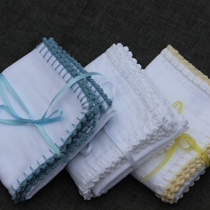 May include: Three folded white handkerchiefs with crocheted edges in teal, white, and yellow. Each is tied with a matching ribbon. The handkerchiefs are neatly stacked, showcasing the delicate crochet work and soft colours.