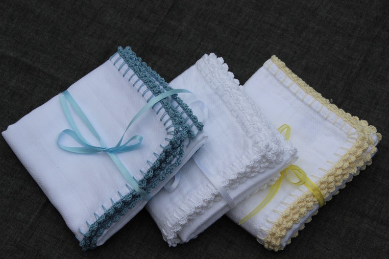 May include: Three folded white handkerchiefs with crocheted edges in teal, white, and yellow. Each is tied with a matching ribbon. The handkerchiefs are neatly stacked, showcasing the delicate crochet work and soft colours.