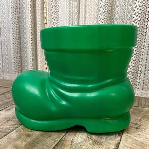 May include: A green plastic boot-shaped planter. The planter is made to look like a boot with a fold at the top.