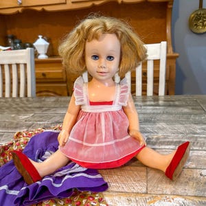May include: A vintage doll with blonde hair and blue eyes, wearing a pink dress with white trim and red shoes. The doll is sitting on a wooden table, with a purple and patterned fabric in the foreground. The doll's dress has a white collar.