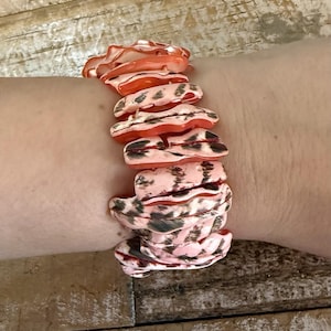 May include: A stretch bracelet composed of irregular, flat, organic-shaped pieces in shades of coral, peach, and light pink. Each piece features dark brown, mottled, or striped patterns across the surface, creating a textured, layered appearance.