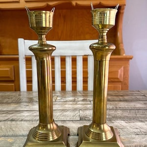 May include: Two antique brass candlesticks. Each candlestick has a square base, a long, cylindrical stem, and a cup-shaped top with a handle. The candlesticks are displayed on a wooden table. The background includes a white chair and wooden cabinets.