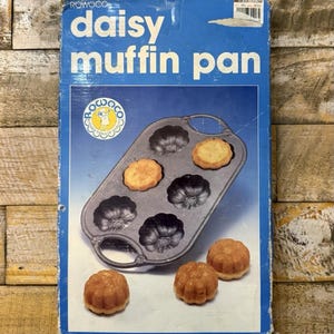 May include: A blue rectangular box for a Rowoco daisy muffin pan. The box features the text daisy muffin pan and a graphic of a dark gray metal pan with six flower-shaped molds, two of which contain baked goods, alongside three baked muffins.