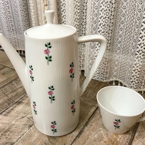 May include: White ceramic teapot and teacup with a floral pattern of pink roses and green leaves. The teapot has a spout and a handle. The teacup has a handle.