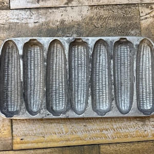 May include: A rectangular cast metal baking pan featuring seven individual corn-shaped molds. The silver-toned pan has a textured surface with detailed kernels and husks in each cavity, and includes two small circular holes on the side tabs for hanging.