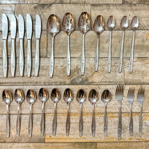 May include: A selection of vintage cutlery, including knives, spoons, serving spoons, and forks, displayed on a wooden surface. The silverware features a floral pattern on the handles and a shiny, metallic finish.