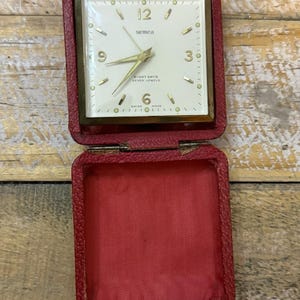 May include: A vintage Semca travel alarm clock in a red leather folding case. The square clock face is white with gold-tone numerals at 3, 6, 9, and 12, gold hands, and text reading Semca, Eight Days, Seven Jewels, and Swiss Made.
