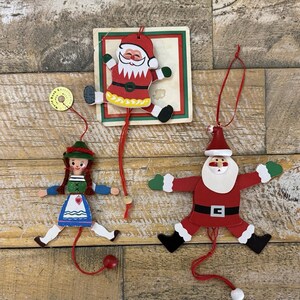 May include: Three wooden jumping jack ornaments featuring a Santa Claus on a square card, a second Santa Claus, and a girl in a traditional dress. Each figure has red strings for movement and painted details in red, white, green, and blue colors.
