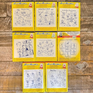 Könnte beinhalten: Acht gelbe Aunt Martha's Bügeltransfer-Packungen mit verschiedenen Strichzeichnungen für Stickereien. Die Titel umfassen Denim Dan, Little Boy's Quilt, Little Boy Tea Towels, Nursery Rhymes, Colonial Girl and Puppy, Mama's Little Helper, Mommy's Little Angel und Saucy Suzy.