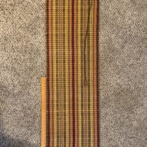 Può includere: Un lungo runner da tavola rettangolare in bambù intrecciato con strisce verticali nei toni del marrone, giallo e arancione. Un bordo in tessuto arancione con un dettaglio a intaglio è fissato in alto. Un righello in legno indica 30,5 cm.