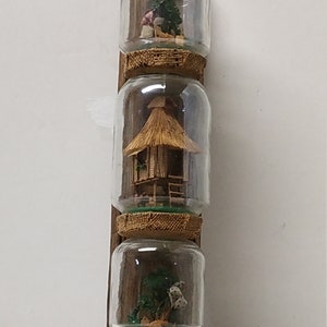 Puede incluir: Tres frascos de vidrio están unidos a una tabla de madera. Cada frasco contiene una escena en miniatura. El frasco superior tiene una persona en miniatura, el frasco del medio tiene una cabaña en miniatura y el frasco inferior tiene una palmera en miniatura.