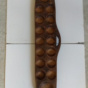 Puede incluir: Un tablero de juego de madera con 16 depresiones redondas, probablemente utilizado para un juego tradicional como el Mancala. El tablero es marrón y tiene un asa tallada.