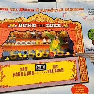 Könnte beinhalten: Eine Karnevals-Spielbox mit einer bunten Illustration eines Enten-Themaspiels. Das Spiel beinhaltet eine Spielzeugpistole und eine digitale Punkteanzeige. Die Box trägt die Aufschrift "Dunk the Duck Carnival Game" und zeigt die Texte "Try Your Luck" und "Hit the Duck".