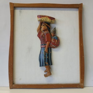 Puede incluir: Una obra de arte mural de madera tallada que representa a una persona que lleva una cesta en la cabeza y a un niño en la espalda. Las figuras visten ropa tradicional colorida. La obra está enmarcada en un marco de madera marrón claro y montada sobre un fondo blanco.