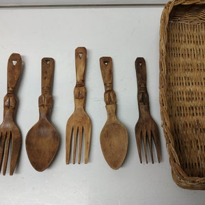 Puede incluir: Un conjunto de seis utensilios de madera tallados a mano, que incluyen cucharas y tenedores, con mangos decorativos. Los utensilios se muestran junto a una cesta de mimbre tejida. La madera tiene un tono cálido y natural.