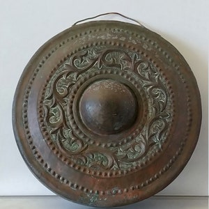 Könnte beinhalten: Ein runder, kupferner Gong mit einem komplizierten, geprägten Blumenmuster. Der Gong hat eine erhöhte Mitte und eine Aufhängeschlaufe oben.