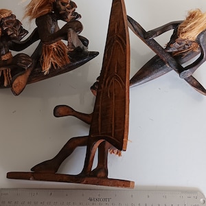 May include: Three wooden carvings of people surfing. The carvings are painted brown and have a rustic look. The carvings are on a white surface with a ruler for scale.